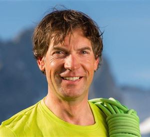 A sporty man with short hair smiles at the camera. He is wearing a green shirt and is holding a climbing rope in his hand, while the mountains are visible in the background.
