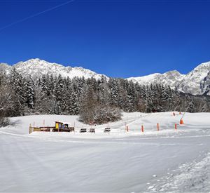 A snowy landscape with high mountains in the background. Some shoveling and seating areas are visible, surrounded by snow-covered trees.