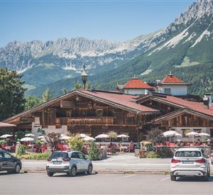 Ein traditionelles Chalet in den Bergen mit blühenden Blumen und Außensitzplätzen. Im Hintergrund sind majestätische Berge und ein klarer blauer Himmel zu sehen.