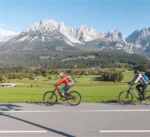 Two cyclists ride along a road on a vast green meadow. In the background, impressive mountains and a clear blue sky can be seen.