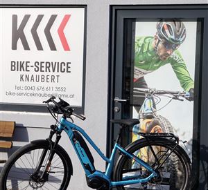 A blue e-bike is standing in front of a shop named "Bike-Service Knaubert". In the background, there is a large image of a cyclist.