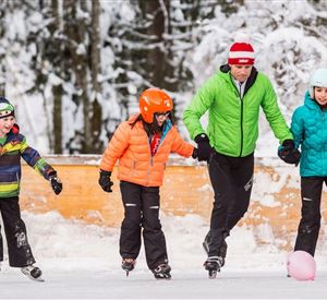 Een vrolijke groep kinderen en een volwassene die aan het schaatsen zijn op een ijsbaan in de sneeuw. Ze dragen kleurrijke winterkleding en hebben veel plezier met samen glijden.