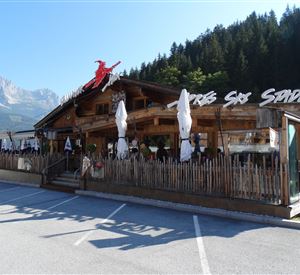Ein traditionelles Restaurant in einer malerischen Umgebung mit Holzverkleidung und Sonnenschirmen. Im Hintergrund sind Bäume und Berge sichtbar.