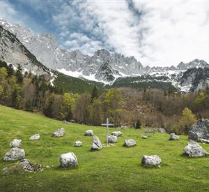A green meadow with a stone circle and a wooden cross in the foreground. In the background, majestic mountains rise up under a cloudy sky.