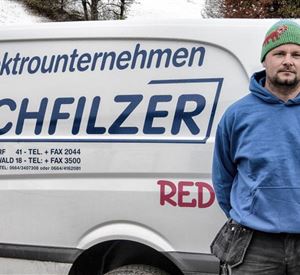 Een vakman staat voor een bestelwagen met de tekst "Elektroniebedrijf Hochfilzer". De man draagt een blauwe hoodie en een groene muts.