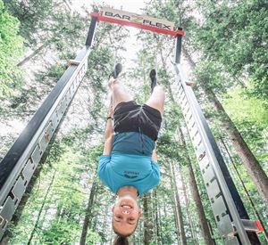 Een jonge man hangt ondersteboven aan een trainingsapparaat in het bos. De omgeving is groen en lichtdoorlatend.