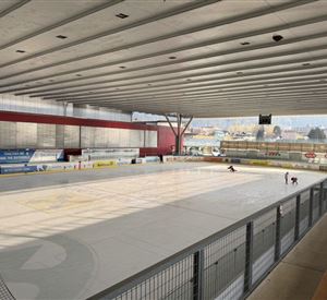 Een ijsbaan met een lege ijsvloer en hoge plafonds. Op de achtergrond zijn enkele skaters zichtbaar.