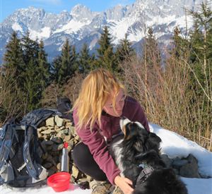 Ein Mann sitzt auf einem Weg und streichelt seinen Hund. Im Hintergrund sind hohe Berge und ein grünes Tal zu sehen.