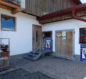 Een gebouw met houten bekleding en twee deuren. Voor de ingangstrap staan planten en posters.