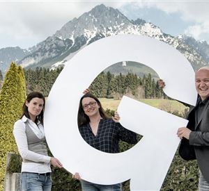 Drie personen houden een grote letter "G" voor een indrukwekkend berglandschap. Op de achtergrond zijn besneeuwde bergen en groene bomen zichtbaar.
