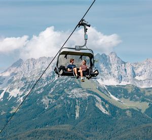 Zwei Personen in einem Sessellift vor dem Bergmassiv des Wilden Kaisers unter blauem Himmel.