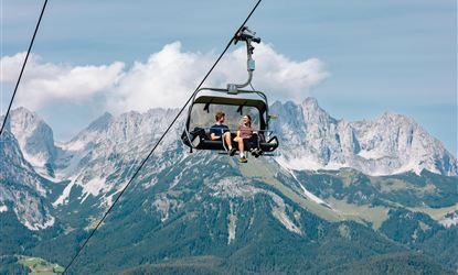 Zwei Personen sitzen in einer Gondel auf dem Weg über den Bergen. Die Landschaft besteht aus grünen Wäldern und majestätischen Gipfeln unter einem klaren Himmel.