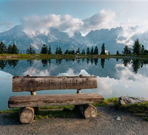 A wooden bench stands by the shore of a calm lake, surrounded by mountains. The sky is partly cloudy and reflects in the water.