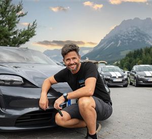 Een man poseert glimlachend naast een modern auto in een bergachtig landschap. 
Achtergrond zijn verdere auto's en een mooie lucht te zien.