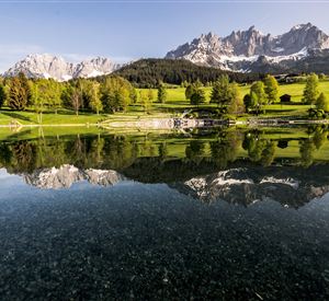 Eine malerische Landschaft mit einem ruhigen See und sanften Hügeln. Im Hintergrund erheben sich majestätische Berge unter einem klaren Himmel.