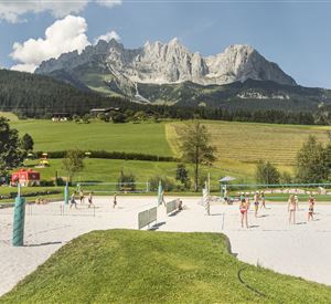 Een zandveld met volleybalspelers voor indrukwekkende bergen. De omgeving is groen en zonnig, ideaal voor sportieve activiteiten.