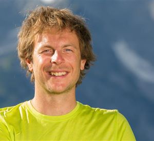 Een glimlachende man draagt een felgroen shirt. Op de achtergrond zijn vage bergen te zien.