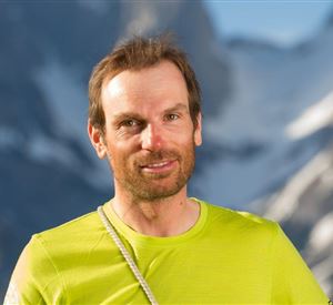 Een man staat voor een bergachtige achtergrond. Hij draagt een lichtgroen sportshirt en heeft een vriendelijke uitstraling.