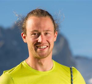 Ein lächelnder Mann in einem leuchtend gelben Shirt. Im Hintergrund sind Berge mit schneebedeckten Gipfeln zu sehen.