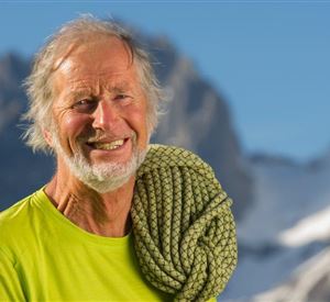 Een lachende oudere man draagt een klimtouw en staat voor een indrukwekkend berglandschap. De lucht is helder en de omgeving ziet er majestueus uit.