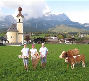 Eine malerische ländliche Szene mit drei Personen in traditioneller Kleidung, die vor einer Kirche stehen. Im Vordergrund sind zwei Kühe auf einer grünen Wiese zu sehen, umgeben von Bergen.