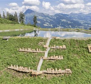 A picturesque landscape with a wooden lounging area amidst green meadows and a tranquil pond. In the background, majestic mountains and a blue sky can be seen.