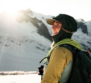 Eine Person steht am Berg und schaut lächelnd in die Ferne. Die Landschaft ist schneebedeckt und die Sonne scheint hell.