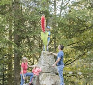 Eine Familie klettert an einem großen Stein im Wald. Ein Kind steht oben auf dem Stein und hält ein buntes Schild.