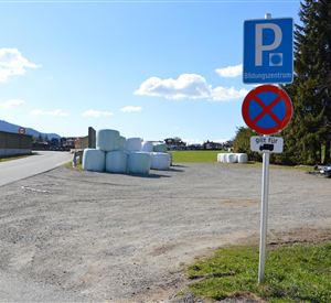 Ein Parkplatz mit einem Parkverbotsschild und runden Ballen im Hintergrund. Der Himmel ist blau mit einigen Wolken.