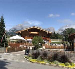 Ein traditionelles Holzhaus mit einem schönen Garten und Bergen im Hintergrund. Der Himmel ist klar und sonnig.