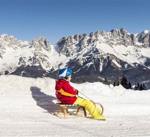 Ein Kind sitzt auf einem Schlitten im Schnee. Im Hintergrund sind beeindruckende Berge zu sehen.