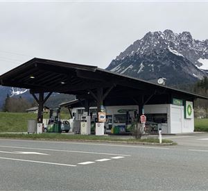 Eine Tankstelle mit einem überdachten Bereich in einer ländlichen Umgebung. Im Hintergrund sind beeindruckende Berge zu sehen.