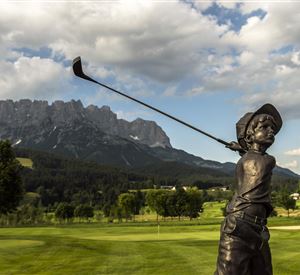 A bronze sculpture of a young golfer stands on a golf course. In the background, majestic mountains and a clear sky can be seen.