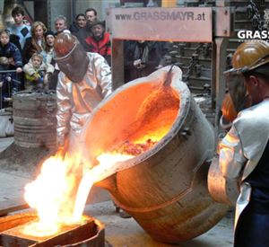 Two people in protective gear are pouring molten metal from a large cauldron. In the background, spectators are watching the spectacle.