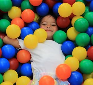 A smiling child is lying in a colorful ball pit. They are surrounded by red, yellow, blue, and green balls.