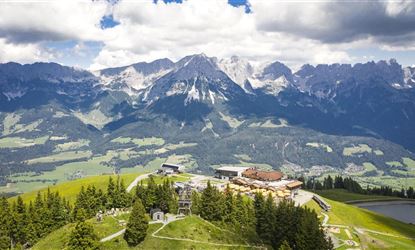 Eine beeindruckende Berglandschaft mit hohen Gipfeln und grünen Wiesen. Im Vordergrund sind Bäume und Gebäude zu sehen, die die natürliche Schönheit der Umgebung ergänzen.
