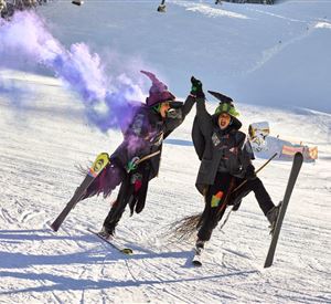 Zwei Personen auf Skiern tragen Hexenkostüme und feiern fröhlich. Sie halten die Hände hoch und lassen lila Rauch steigen.