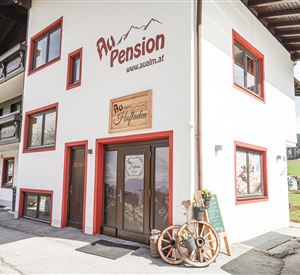 Een gezellige pension met rode raamomlijsting en een uitnodigende ingang. Op de achtergrond zijn groene heuvels en bossen te zien.