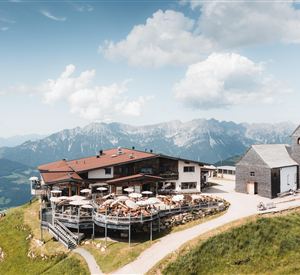 A picturesque mountain cabin with terraces and views of the surrounding mountains. The sky is clear and sunny, ideal for a glimpse into nature.
