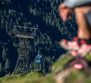 Eine Seilbahn fährt über eine grüne Berglandschaft. Im Vordergrund befindet sich eine Person, die auf dem Boden sitzt.