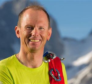 A smiling man in a bright yellow shirt stands in front of a mountainous landscape. He is wearing climbing gear and radiates a sense of adventure.