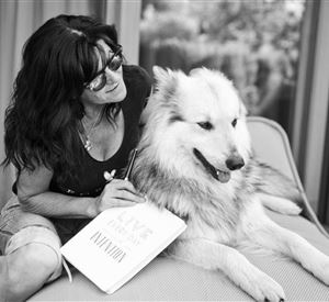 Eine Frau sitzt auf einer Liege und streichelt ihren Hund. Sie hält ein Notizbuch in der Hand und lächelt freundlich.