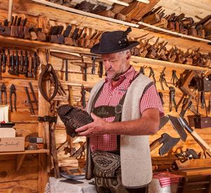 A craftsman in traditional clothing is examining an intricately carved wooden piece. In the background, many tools are visible on a wooden wall.