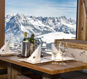 Ein elegantes Restaurant mit einem Holztisch und festlich eingedecktem Geschirr. Im Hintergrund ist eine beeindruckende Berglandschaft mit Schnee zu sehen.