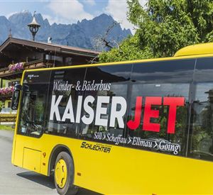 A yellow bus with the inscription "KaiserJET" drives through a picturesque landscape. In the background, mountains and a cozy alpine hut can be seen.