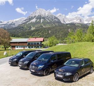 Eine Gruppe von Fahrzeugen steht vor einem malerischen Bergpanorama. Im Hintergrund sind grüne Wiesen und schneebedeckte Gipfel zu sehen.