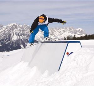 Ein Snowboarder springt über ein Hindernis in einer verschneiten Berglandschaft. Im Hintergrund sind mächtige Berge und ein blauer Himmel zu sehen.
