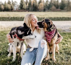 Een vrouw zit in het gras en omarmt twee honden. Ze glimlacht gelukkig terwijl de honden liefdevol bij haar zijn.