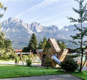 Een speelplaatslandschap met een creatief houten speeltoestel en schommels, omringd door groene weiden. Op de achtergrond stijgen majestueuze bergen op onder een heldere lucht.