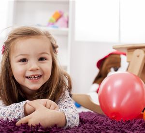 A cheerful girl is lying on a purple carpet and smiling. Colorful toys and a red ball are around her.